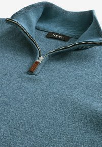 Pull-over en maille bleue avec une fermeture éclair métallique à quart de tour dotée d'une languette en cuir marron et un col haut côtelé, étiquette de marque "NEXT" à l'intérieur.