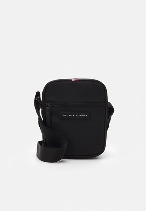 Sac bandoulière noir avec sangle réglable, poche avant zippée et patch logo Tommy Hilfiger sur le panneau avant.