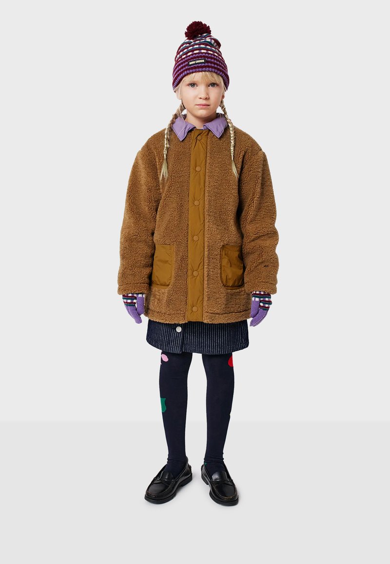 Braune Fleecejacke mit Knopfverschluss und Vordertaschen, kombiniert mit einem lila Hemd mit Kragen, einem gestreiften Hut, Handschuhen und gemusterten Strumpfhosen.