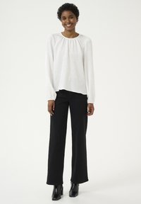 Blouse blanche texturée à manches longues et encolure froncée, associée à un jean noir taille haute à jambes larges et des bottines noires.