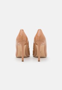 Un paio di scarpe con tacco alto beige lucido viste da dietro, evidenziando i tacchi a punta e la finitura elegante su uno sfondo bianco.