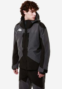 Giacca isolante da uomo in nero e grigio, con cappuccio, tasche con cerniera e un design geometrico con textures e cuciture a contrasto.