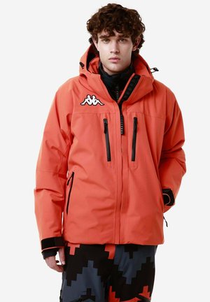 Giacca impermeabile arancione con cappuccio, dotata di tasche con zip nere e un logo bianco sul petto. Indossata sopra pantaloni a fantasia.