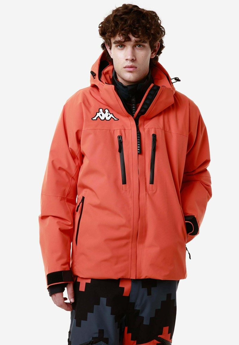 Giacca impermeabile arancione con cappuccio, dotata di tasche con zip nere e un logo bianco sul petto. Indossata sopra pantaloni a fantasia.