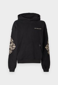 Melnā hoody ar priekšējo kabatu, zelta teksts "NEIZTIEK KULTŪRU" un izsmalcināts zelta raksts uz piedurknēm. Mīksts audums un brīvs piegriezums.