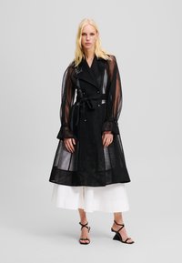 Sort mesh trenchcoat med lange ærmer, bælte i taljen og udskårne manchetter, lagdelt over en hvid nederdel. Har for- og bagknapper samt et signaturdetalje.