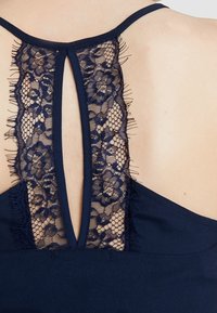 Vêtement bleu marine vu de dos avec une ouverture en forme de clé verticale bordée de dentelle florale et de fines bretelles sur une peau claire.