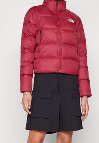 Mujer vestida con una chaqueta de plumas roja corta y una falda cargo negra con grandes bolsillos frontales, de pie contra un fondo blanco.