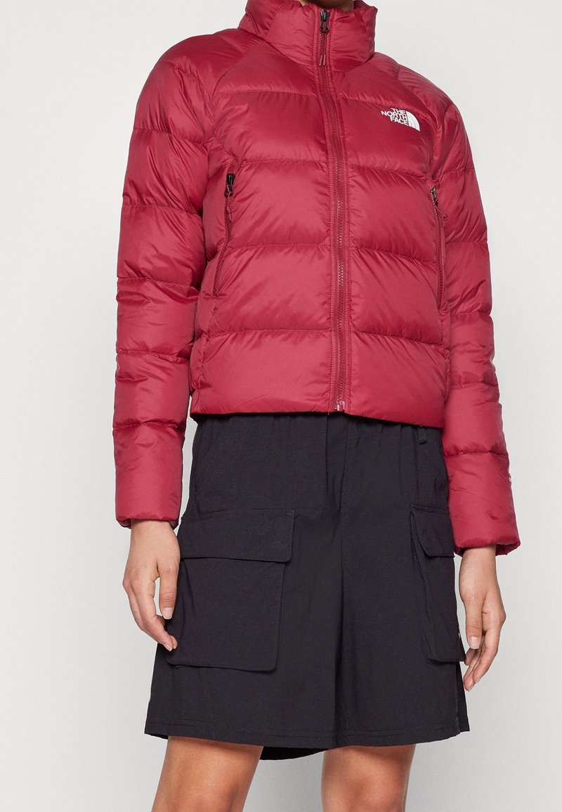Mujer vestida con una chaqueta de plumas roja corta y una falda cargo negra con grandes bolsillos frontales, de pie contra un fondo blanco.