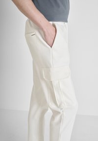 Weiße Cargo-Hosen mit glatter Textur, ausgestattet mit Seitentaschen und dezenten Akzenten. Entworfen mit einer lockeren Passform und elastischem Bund.