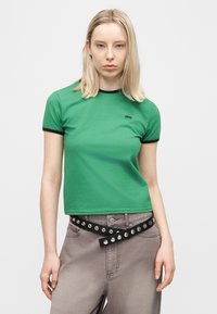 Νέα γυναίκα με ίσια ξανθά μαλλιά, φορώντας πράσινο T-shirt με μαύρο τελείωμα και γκρι παντελόνι με μαύρη ζώνη με τρουκς.