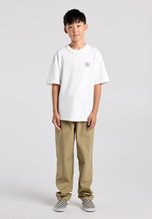 Jongen staat in beige broek, wit oversized T-shirt met klein logo op de borst, en zwart-witte geruite slip-on schoenen tegen een effen achtergrond.