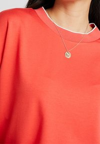 Sudadera coral con un borde de cuello blanco a contraste, de corte relajado y textura suave, combinada con un delicado collar de oro.