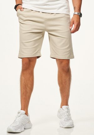 Mann trägt beige Shorts, weiße Sneakers und weißes T-Shirt, steht mit einer Hand in der Tasche vor einem einfarbigen Hintergrund.