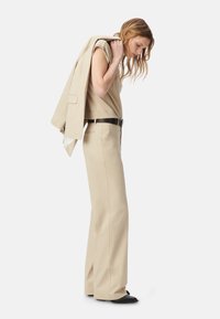 Beige op maat gemaakte broek met een krijtstreep patroon, gecombineerd met een bijpassende mouwloze top. Een lichte jas over één schouder gedrapeerd. Zwarte riem en schoenen.