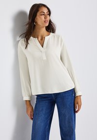 Femme portant un haut à manches longues blanc cassé avec un décolleté fendu et un jean bleu, se tenant devant un mur blanc.