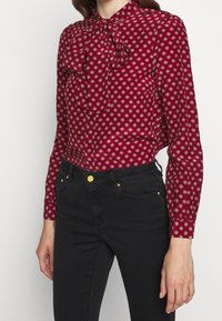 Mujer vestida con una blusa roja de manga larga, con estampado y un lazo en el cuello, combinada con unos vaqueros negros de cintura alta que presentan botones y remaches dorados.