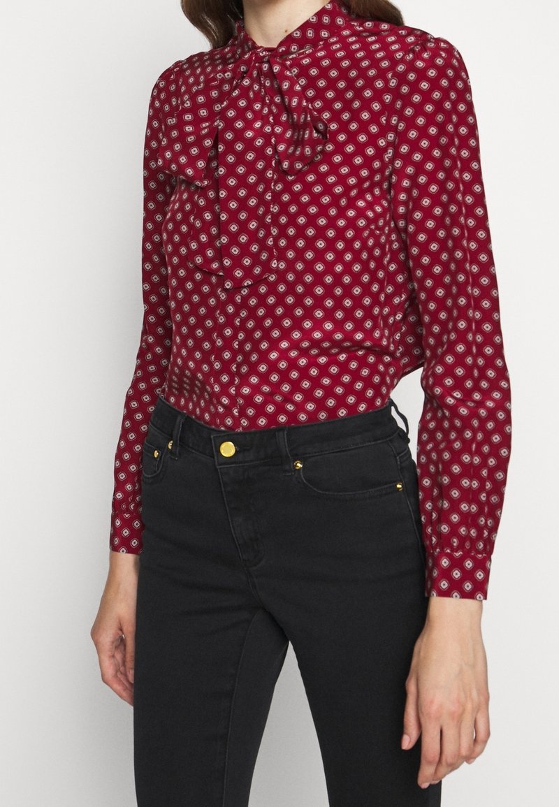 Mujer vestida con una blusa roja de manga larga, con estampado y un lazo en el cuello, combinada con unos vaqueros negros de cintura alta que presentan botones y remaches dorados.