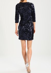 Abito blu scuro scintillante con paillettes, maniche a tre quarti, silhouette aderente e leggero design a incrocio. Sandali argento con tacco alto completano il look.