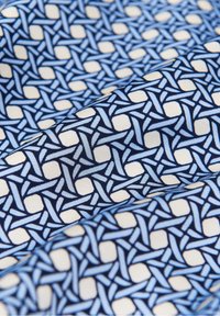 Mönstrad tyg med sammanflätade blå och ljusblå geometriska mönster på en krämfärgad bakgrund. Mjuk textur med en slät finish.