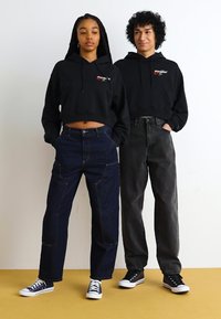 To personer har på seg svarte hettegensere med et logo, kombinert med mørke denimjeans. Klærne har kontrastfarger og avslappede joggesko.