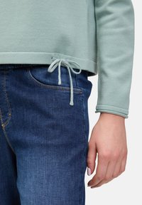 Jasnozielona bluza z kapturem, z detalem sznurka na dole, zestawiona z ciemnoniebieskimi dżinsami z małą kieszonką z przodu.
