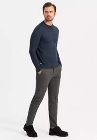 Marineblauer Pullover mit rundem Halsausschnitt, kombiniert mit dunkelgrauen Slim-Fit-Hosen. Das Model steht mit den Händen in den Taschen und trägt schwarze Schuhe.