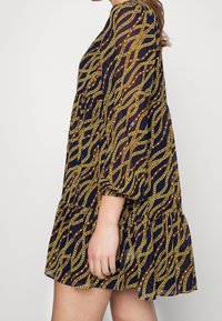 Pitkähihainen mekko, jossa on tummansininen tausta ja kultaketju-kuvio, kerroksellinen helma sekä kevyt, ilmava kangas.