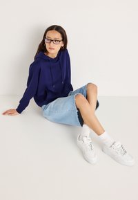 Tmavomodrá mikina s kapucňou, svetlomodrá džínsová sukňa s rozstrapkaným lemom, biele tenisky a biele ponožky po členky, sediaca na jednoduchom pozadí.