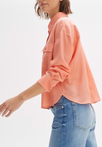 Licht koraalkleurig, langstaand shirt met een kraag, voorzien van een borstzak en een gebogen zoom, gemaakt van zachte stof, gecombineerd met blauwe jeans.