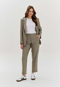 Kobieta z długimi brązowymi włosami uśmiecha się, ubrana w beżowy, w kratkę garnitur, biały top oraz białe sneakersy z czarnymi paskami, stojąc na jednolitym tle.