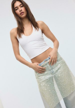 Frau trägt ein ärmelloses weißes Crop-Top und hellblaue Jeans mit einem transparenten, mit Pailletten besetzten Rocküberzug, sie richtet den Taillenbund mit beiden Händen.