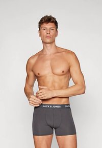 Póló nélküli férfi, rövid göndör hajjal, sötétszürke Jack & Jones boxer alsóneműben, sima világos háttér előtt áll.