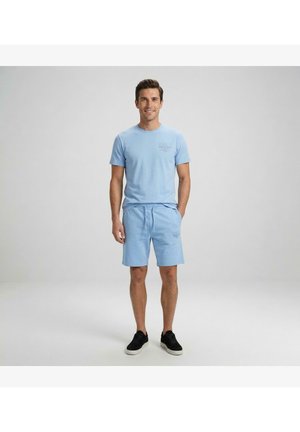 Man staand, draagt een bijpassend lichtblauw T-shirt en korte broek met zwarte sneakers, glimlachend tegen een effen grijze achtergrond.
