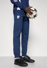 Navy sweatpants with cuffed ankles, featuring a logo patch. Black athletic shoes with a textured design and shin guards. Soccer ball held in hands.