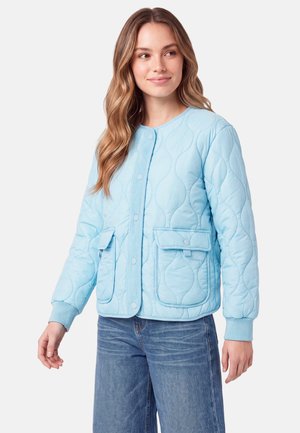 Frau mit welligem Haar, die eine hellblaue gesteppte Jacke mit Druckknöpfen und zwei vorderen Klappentaschen trägt, kombiniert mit blauen Jeans.