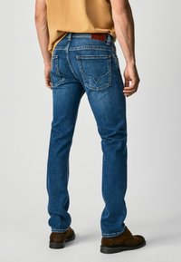 Blue denim jeans with a slim fit, featuring back pockets with distinctive stitching, a leather patch on the waistband, and a clean hem.
