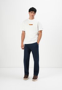 Young man wearing white Tommy Hilfiger T-shirt, dark blue pants, and brown sneakers standing with one hand in pocket against plain white background.