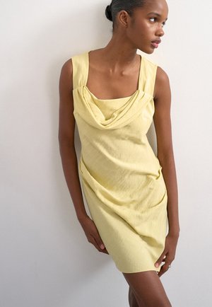 Mujer de piel oscura con un vestido amarillo sin mangas y escote drapeado, de pie de lado contra una pared blanca lisa.