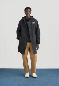 Μαύρο αδιάβροχο παρκά με κουκούλα, κουμπιά με κούμπωμα, μεγάλες μπροστινές τσέπες και λογότυπο στο στήθος, συνδυασμένο με καφέ παντελόνια και λευκά αθλητικά παπούτσια.