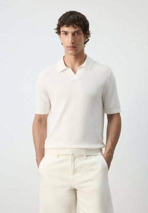 Hombre con pelo corto y rizado, vistiendo una camisa polo blanca de manga corta con textura y pantalones blancos, de pie con las manos en los bolsillos sobre un fondo liso.