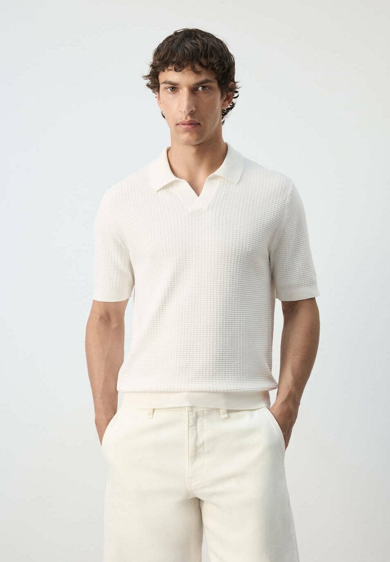 Homme aux cheveux courts et bouclés portant un polo blanc texturé à manches courtes et un pantalon blanc, debout les mains dans les poches devant un fond uni.