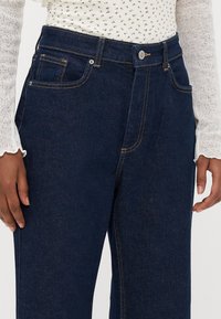 Dark blue denim jeans with a straight cut, featuring visible stitching, a front button, and multiple pockets. Soft textured fabric.
