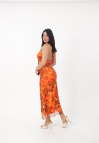 Oranje bloemen maxi jurk met een rugloze ontwerp, lichtgewicht stof en een zijsplit. Gecombineerd met witte hoge hakken sandalen.