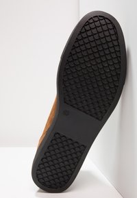 Brązowy zamszowy but z teksturowaną czarną gumową podeszwą z wzorem diamentowym i logo, ustawiony pod kątem na neutralnym tle.