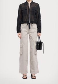 Chaqueta negra transparente con cremallera y dos bolsillos en el pecho, combinada con pantalones cargo gris claro de diseño multi-bolsillo. Un bolso negro y sandalias completan el look.