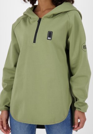 Olijfgroene capuchontrui met zwarte halve rits en logopatch op de borst en linker bovenarm, gedragen met een blauwe spijkerbroek.