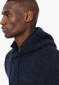 Profilo laterale di un uomo che indossa un hoodie blu scuro con un piccolo logo ricamato sul petto, che guarda avanti contro uno sfondo bianco.