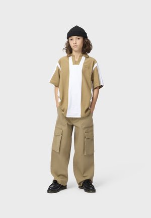 Ung person iført en beige og hvid oversized skjorte, beige cargobukser, sorte sko og en sort beanie, stående med hænderne i lommerne.