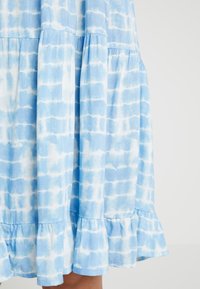 Blaues Baumwollkleid mit einem Batikmuster in hellen und dunklen Blautönen, mit vollem Saum und geschichteter Gestaltung. Weiche Textur und fließende Silhouette.